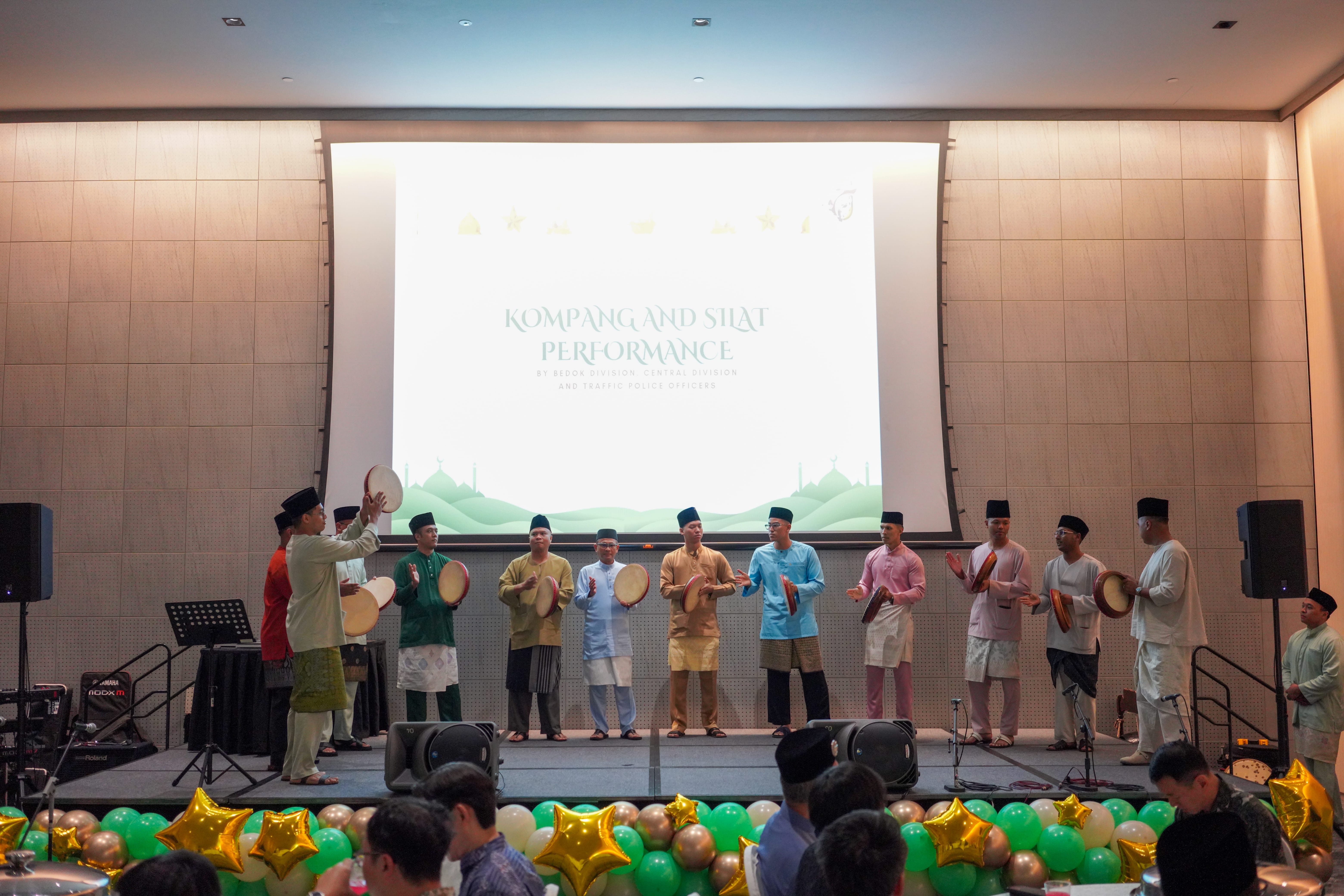 A kompang and silat group performing on stage, with musicians holding frame drums in front of a large projection screen.